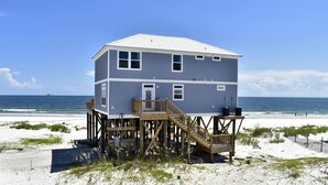 A two-story beach house with a wooden deck and stairs, located on a sandy beach with the ocean in the background. - Island Time IV - 101 Pushmatah (Dauphin Island)
