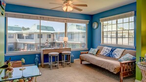 Ein Zimmer mit blauer Wand, Deckenventilator, einer Couch, einem Couchtisch, einem Schreibtisch mit zwei Hocker und großen Fenstern mit Blick auf Häuser.