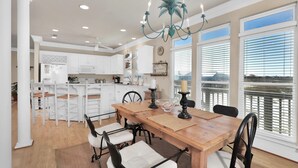 A spacious dining area with a wooden table and chairs, a chandelier, and large windows with a view of the water. - Tramarlis - 2254 Sandpiper (Dauphin Island)