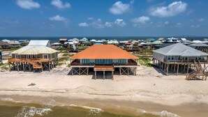 A beachfront property with multiple houses on stilts, a central building with an orange roof, and a view of the ocean and a bridge in the background.