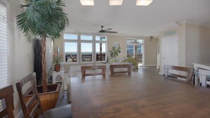 A spacious living area with a large wooden dining table, a ceiling fan, and a view of the ocean through large windows. - Wave Loudest - 103 Pushmata (Dauphin Island)