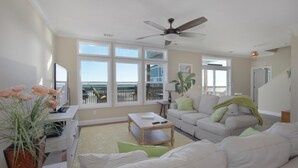 A spacious living room with a large window, a ceiling fan, and a view of the outside. - Wave Loudest - 103 Pushmata (Dauphin Island)