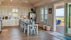 A spacious kitchen with white cabinets, a dining area with white chairs, a large window with blinds, and a view of the beach.