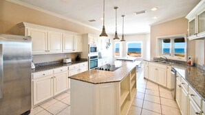 A spacious kitchen with white cabinetry, granite countertops, and a large island. It features a built-in oven, microwave, and a view of the ocean through the windows. - Tidal Wave - 2636 Bienville (Dauphin Island)