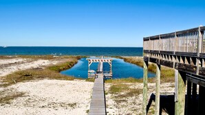 A wooden boardwalk leading to a pier over a body of water, with a sandy beach and grassy areas visible. - Tidal Wave - 2636 Bienville (Dauphin Island)