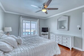 Een slaapkamer met een bed, een plafondventilator, een televisie, een dressoir en een groot raam met uitzicht op de omgeving.