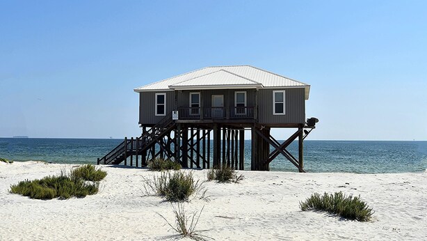 Una casa de madera sobre pilotes con balcón, situada en una playa arenosa y el océano al fondo.