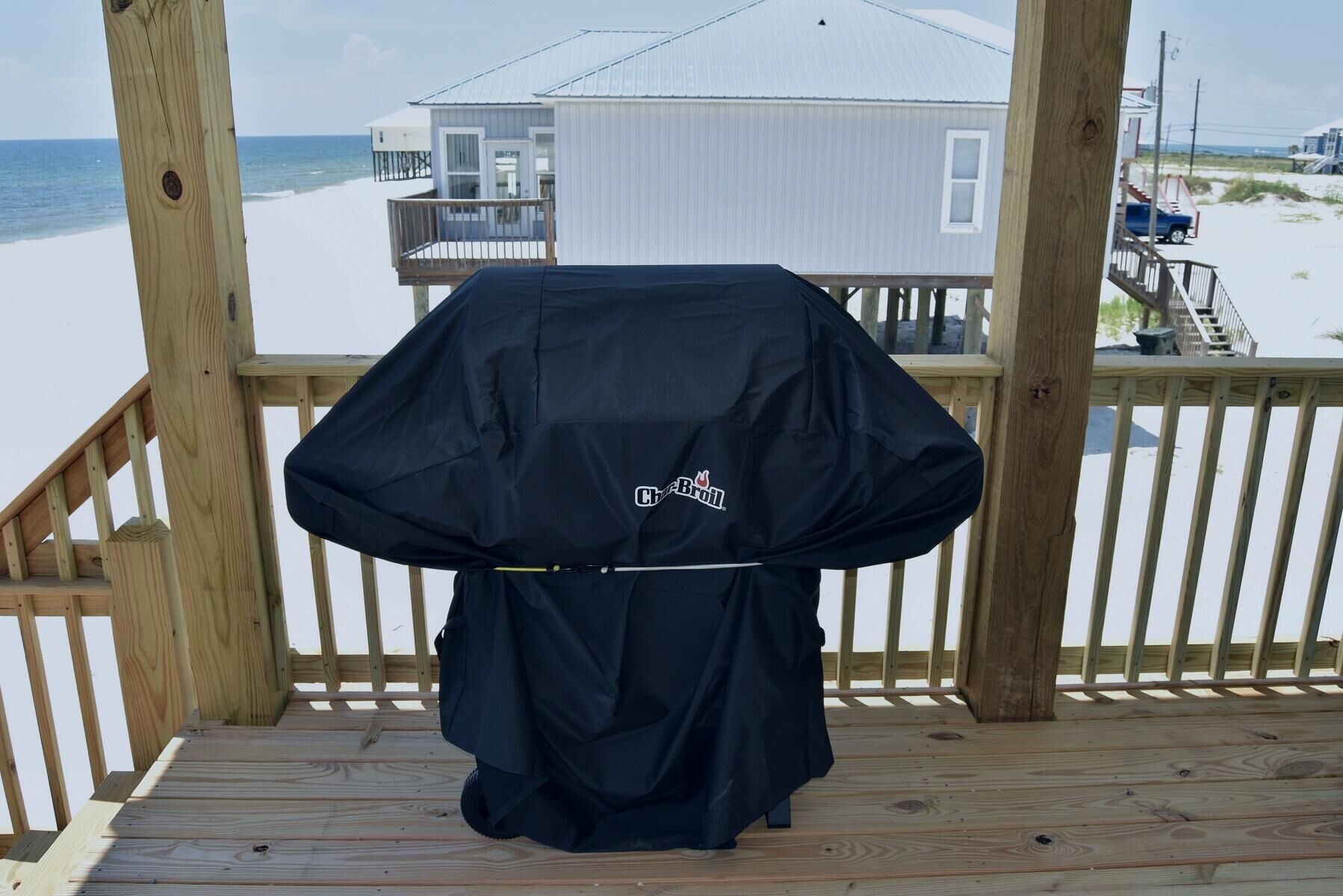 A barbecue grill on a wooden deck overlooking a beach with a house in the background.