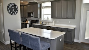 A modern kitchen with grey cabinets, a central island with bar stools, a built-in microwave, and a large wall clock.