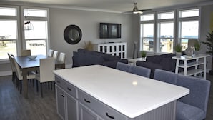 A modern kitchen with a central island, dining area with chairs, and a TV mounted on the wall. - Sonny Daze - 105 Quebec Ct (Dauphin Island)