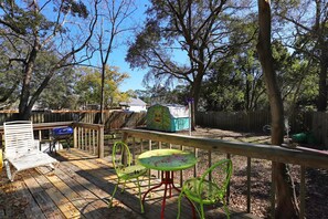 A wooden deck with a table and chairs, surrounded by trees and a fenced yard. - Surf Shack - 1013 Cadillac (Dauphin Island)