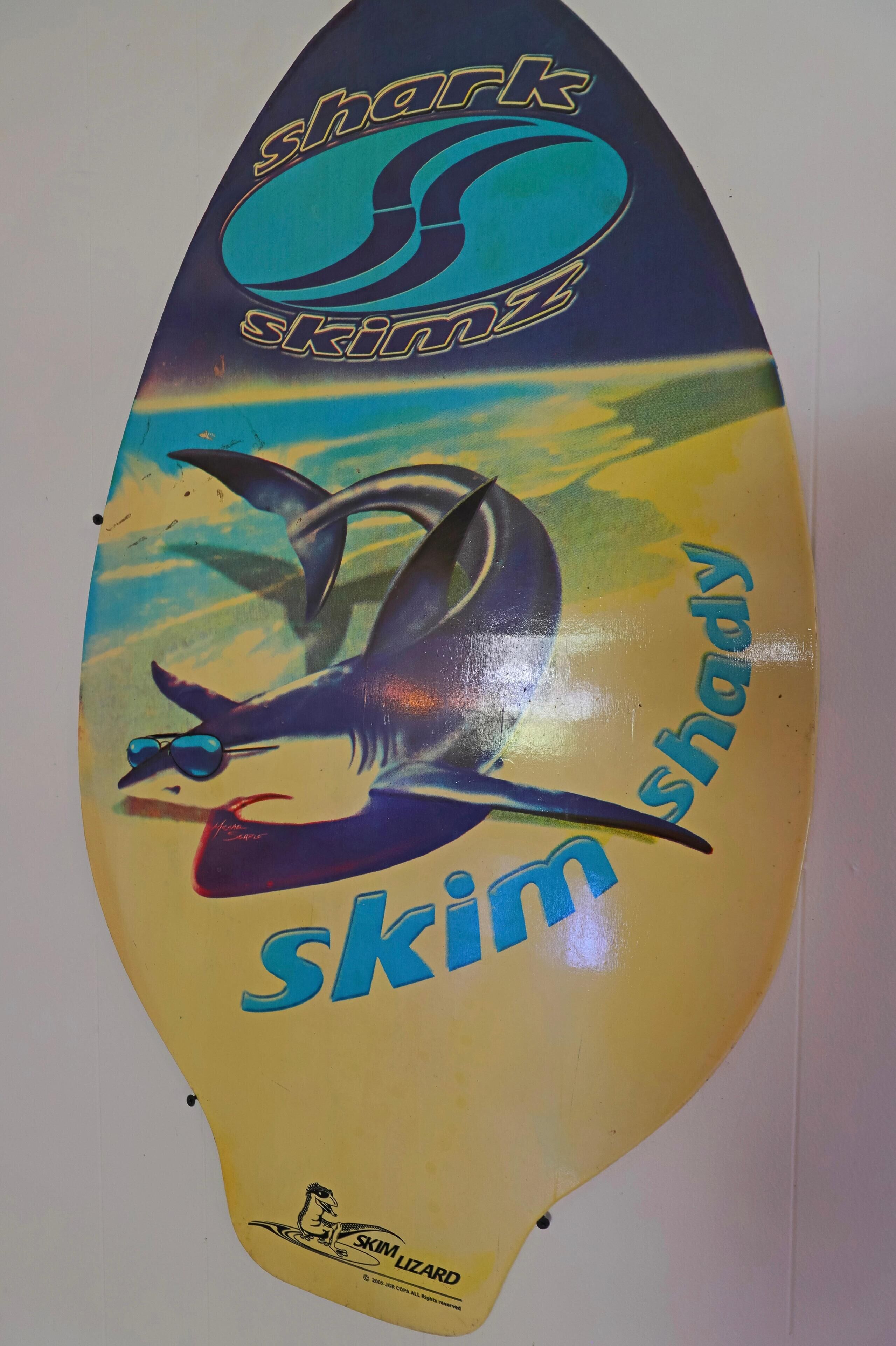 Una tabla de surf con un tiburón y el texto "Shark Skim" y "Skim shady".
