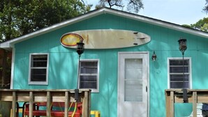 A turquoise house with a white door, two windows, and a surfboard mounted on the wall. - Surf Shack - 1013 Cadillac (Dauphin Island)