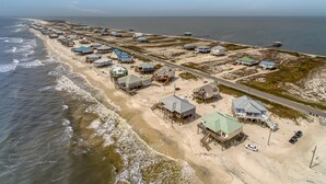 Vista aérea de una zona costera con varias casas de playa, algunas con estructuras elevadas, y una carretera que recorre la playa.