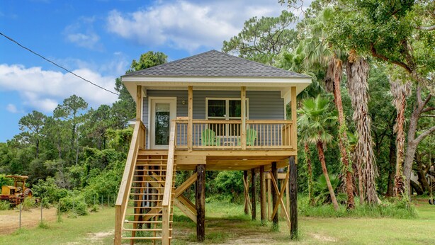 Una casa de madera con terraza y escalera que lleva a un balcón, rodeada de árboles y un cielo despejado.