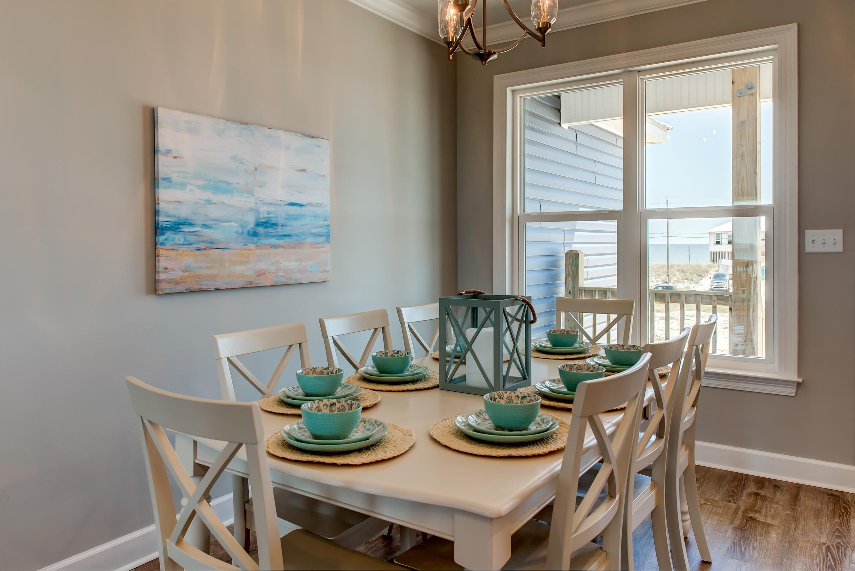 Een eetruimte met een tafel voor zes personen, witte stoelen, turquoise servies en een schilderij met een strandthema aan de muur.