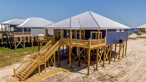 Una casa de playa de madera con un balcón y escaleras, elevada sobre pilotes sobre arena.