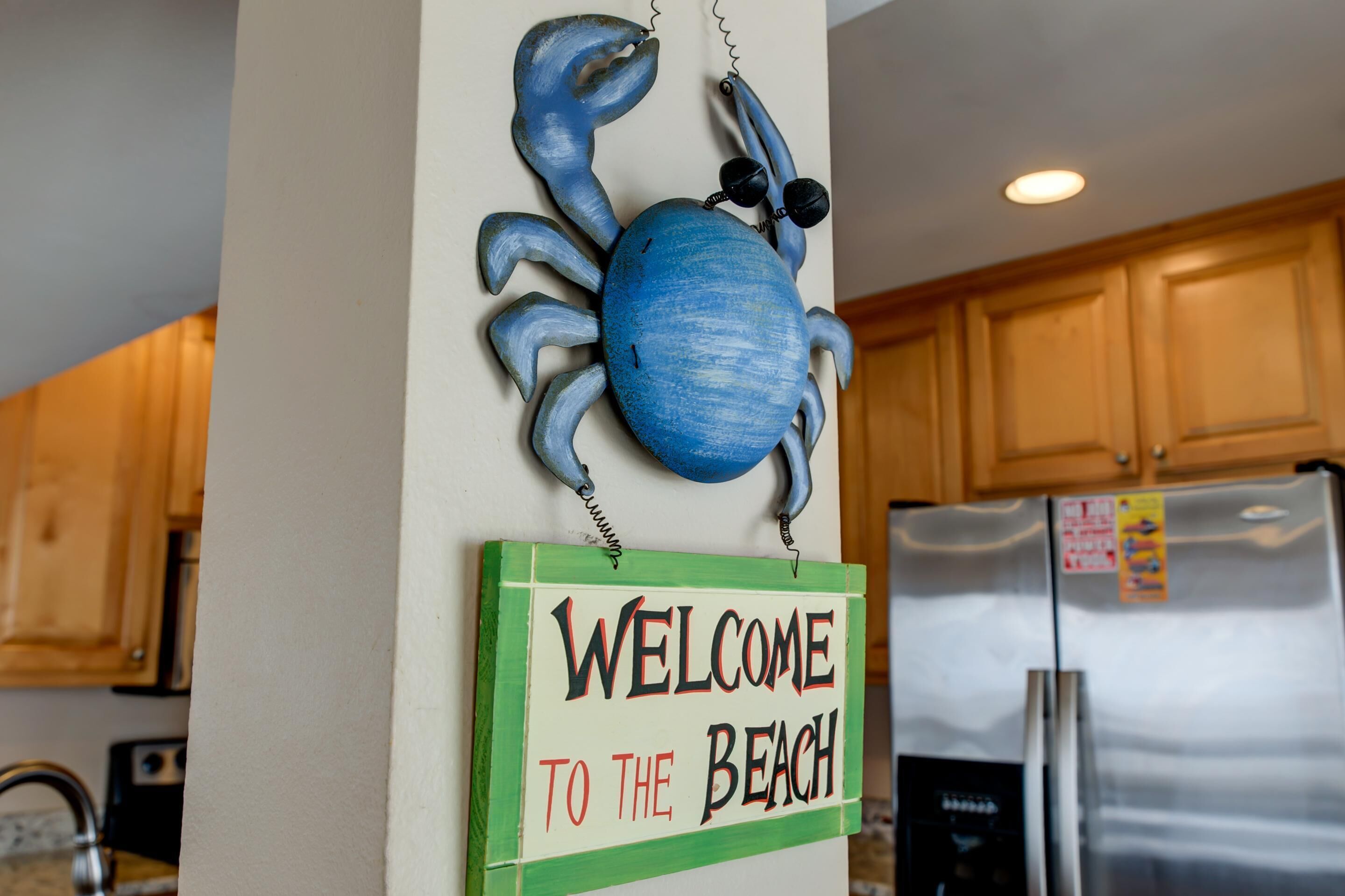 A kitchen with wooden cabinets, a stainless steel refrigerator, a blue crab decoration, and a 'Welcome to the Beach' sign.