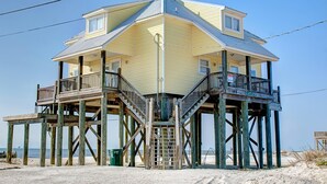 A two-story wooden structure with a balcony and a staircase, situated on a sandy beach.