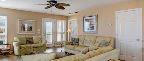 A spacious living room with a large beige sectional sofa, a glass coffee table, and a ceiling fan. There are framed pictures on the walls and a door to the right.