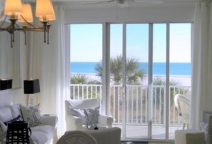 A room with a large sliding glass door leading to a balcony with a view of the ocean. There is a white sofa and a chair inside the room. - Ocean's Edge - Inn #205 (Dauphin Island)