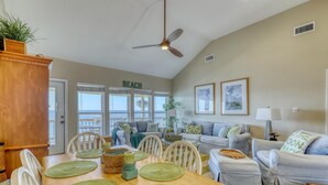 A spacious living room with a dining area, a ceiling fan, and a view of the beach through large windows.