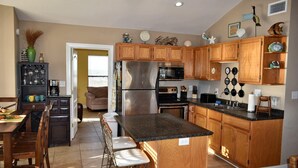 Una cocina con armarios de madera, un refrigerador de acero inoxidable, un microondas y un lavamanos. Hay una zona de comedor con una mesa y sillas.