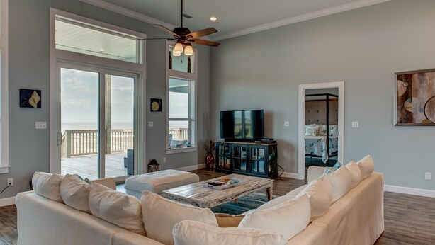 A spacious living room with a large window, a ceiling fan, a flat-screen TV, and a wooden coffee table. - Serenity Now - 2442 Bienville (Dauphin Island)