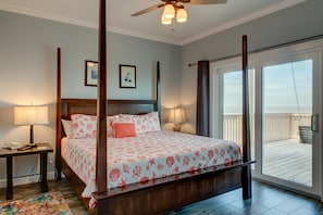 A four-poster bed with a floral bedspread, a nightstand with a lamp, and a view of the ocean through a sliding glass door. - Serenity Now - 2442 Bienville (Dauphin Island)