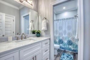 A bathroom with a white vanity, a large mirror, a sink with two faucets, a potted plant, and a shower with a patterned curtain. - Endless Possibilities-1809 Bie (Dauphin Island)