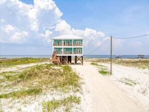 A two-story beach house with a sandy path leading to it, surrounded by dunes and the ocean.