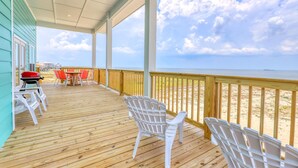 A wooden deck with white plastic chairs and a barbecue grill, overlooking a sandy beach.