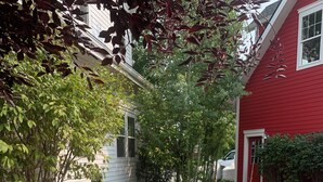 Ein steiniger Weg führt zu einem Haus mit roter und weißer Fassade. Vor dem Haus erstreckt sich eine Rasenfläche und ein Baum mit grünen Blättern.