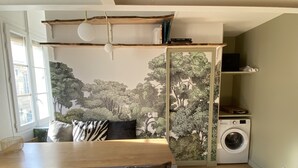 A room with a wooden dining table, chairs, a sofa with patterned cushions, a washing machine, and a bookshelf with books.