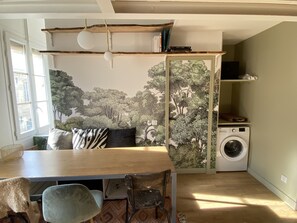 Una habitación con una mesa de comedor de madera, sillas, un sofá con cojines estampados, una lavadora y una estantería llena de libros.
