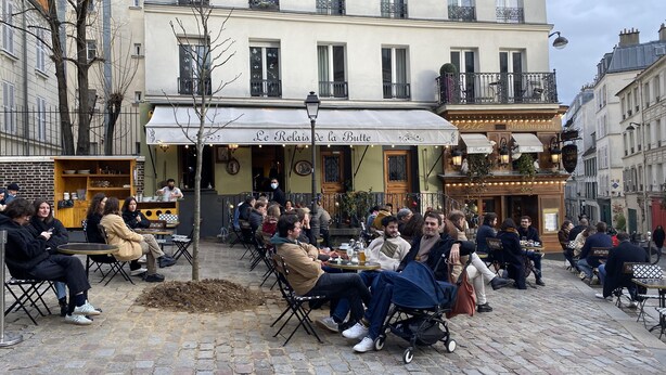 A café with outdoor seating on a cobblestone street, featuring a white awning and a sign reading "Le Relais de la Butte."