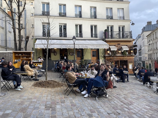 Un café con terraza al aire libre en una calle adoquinada, con un toldo blanco y un letrero que dice "Le Relais de la Butte".