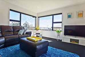 A modern living room with a leather sofa, a blue rug, a coffee table with a tray, a TV, and large windows with a view. - House of Hope - Spa Pool, Sleeps 12 (Rotorua)