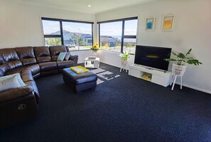 A living room with a brown leather sofa, a white coffee table, a flat-screen TV, and a large window with a view of houses. - House of Hope - Spa Pool, Sleeps 12 (Rotorua)