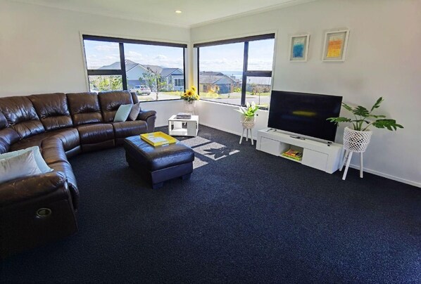 A living room with a brown leather sofa, a white coffee table, a flat-screen TV, and a large window with a view of houses.