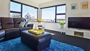 Ein modernes Wohnzimmer mit einer Ledersofa, einem schwarzen Ottomanen, einem Flachbildfernseher und großen Fenstern mit Blick ins Freie.