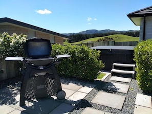 A patio with a Weber grill, a stone pathway, and a view of a house and hills. - House of Hope - Spa Pool, Sleeps 12 (Rotorua)