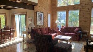 A cozy cabin interior with a wooden floor, a red patterned sofa, a plaid armchair, and a dining table with chairs.