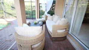 Un espace extérieur aménagé, abrité et doté de mobilier en osier, d’une table avec des tasses et un objet vert, et offrant une vue sur la verdure à travers de grandes fenêtres.