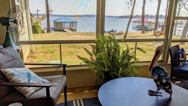 Une pièce avec vue sur un lac, une table à manger, une chaise, un canapé, une plante en pot et une sculpture d’oiseau.