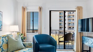 A modern living room with a sofa, armchair, and a glass coffee table. There is a TV on the right and a balcony with chairs. - Gulf Shores Plantation-East 3210 (Gulf Shores)