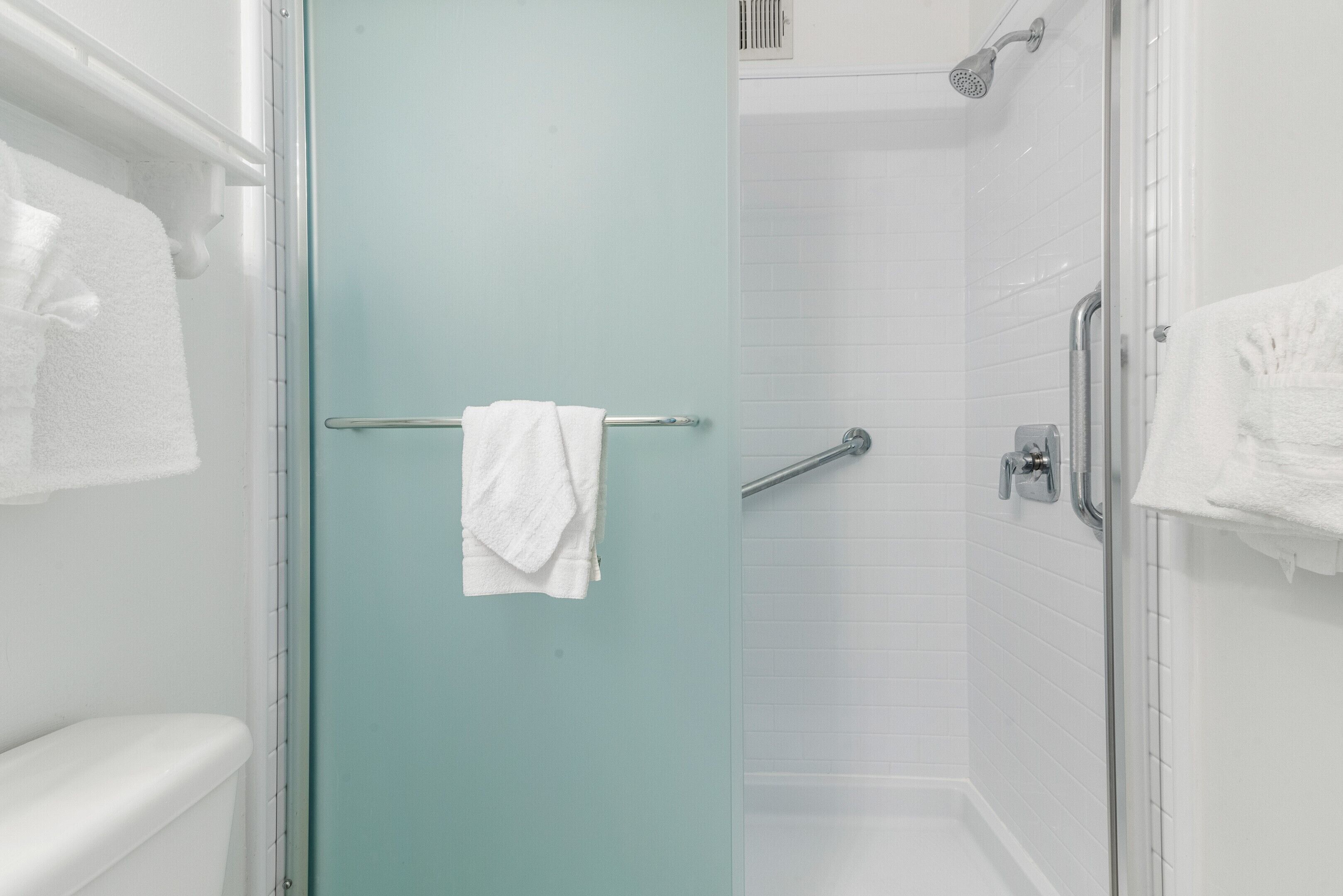 Un baño con mampara de vidrio, paredes con azulejos blancos, un inodoro blanco y un perchero con toalla.