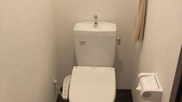 A small, compact bathroom with a white toilet, a paper towel dispenser, and a toilet paper holder on the wall.