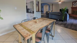 Una zona de menjador amb una taula de fusta i cadires metàl·liques, una cuina al fons i una sala d'estar amb un sofà i una planta.