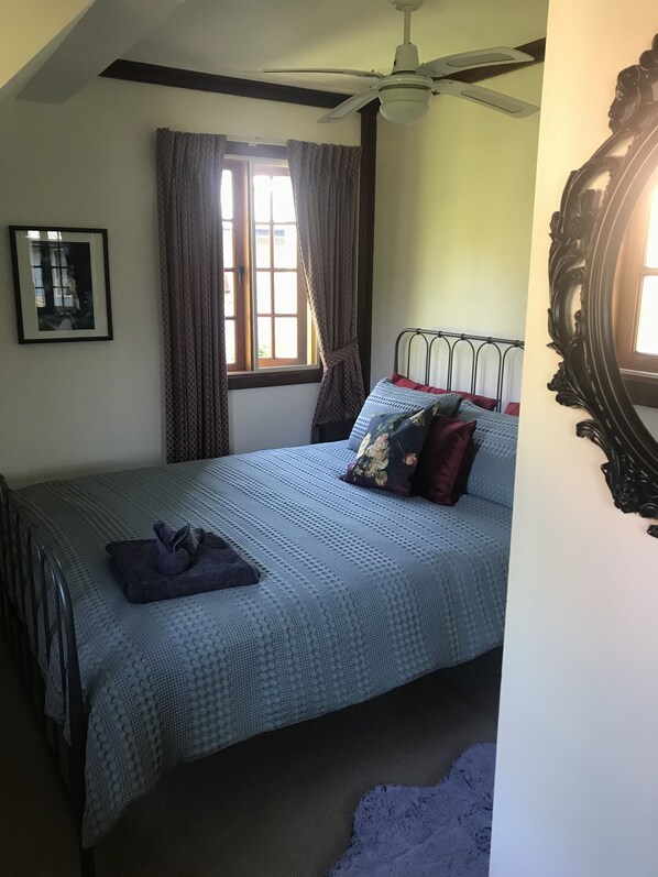 Un dormitorio con una cama, un ventilador de techo, una ventana con cortinas, un cuadro enmarcado y un espejo.